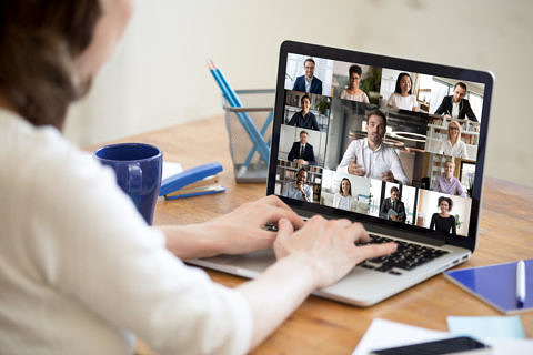 On laptop screen web cam view diverse age and ethnicity businesspeople taking part at group videocall, view over woman shoulder sit at desk working from home. Distant communication, modern app concept